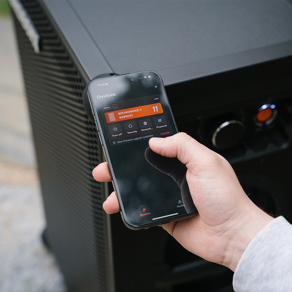 A person holds a smartphone in their hand, displaying the SOUNDBOKS app with options for controlling connected devices. Behind the phone is a SOUNDBOKS Go party speaker, with visible control buttons.